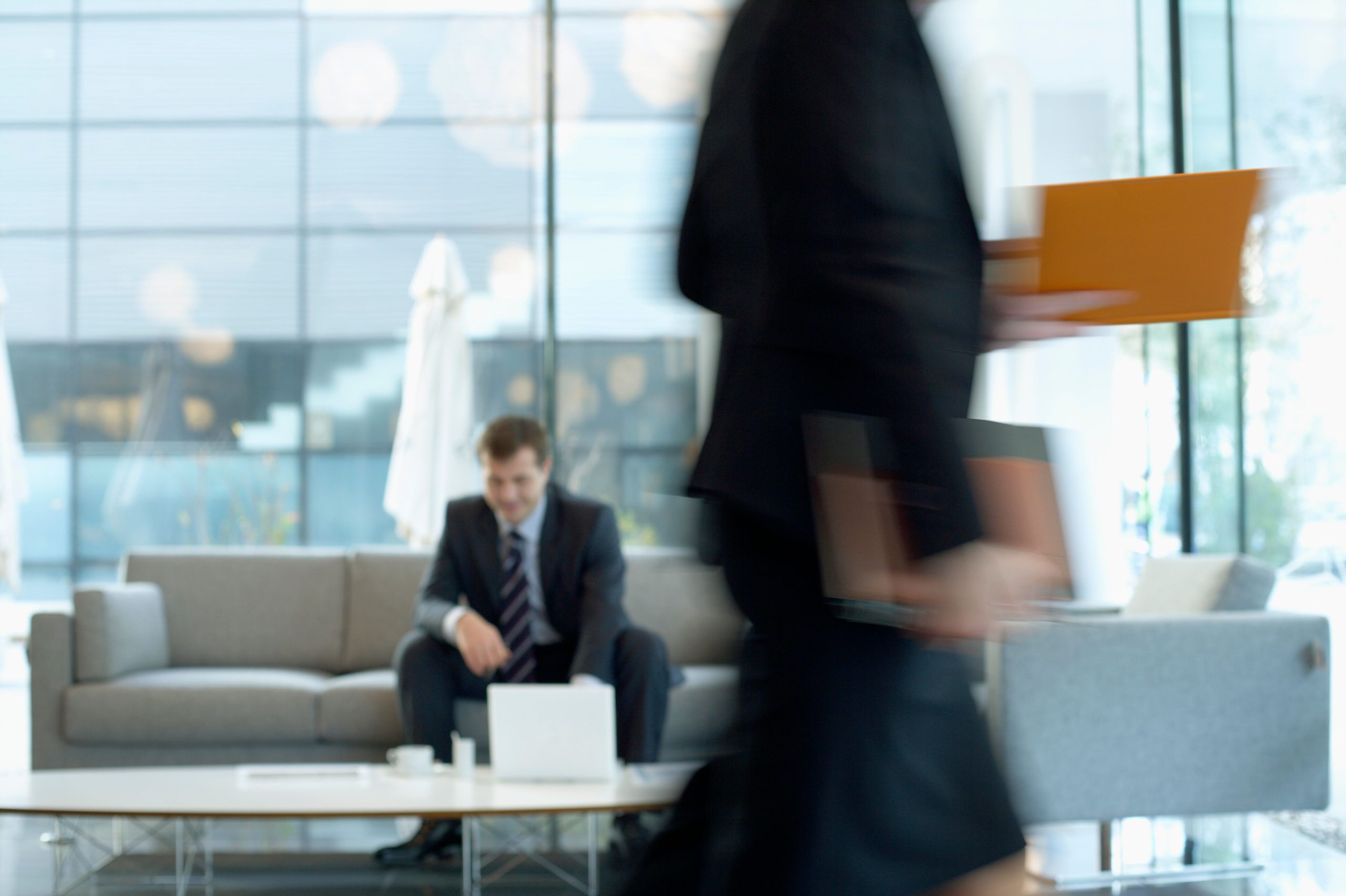 pexels-photo-12585842-12585842 Businessman sitting in modern office lounge using a laptop while colleague walks past, creating dynamic blur.